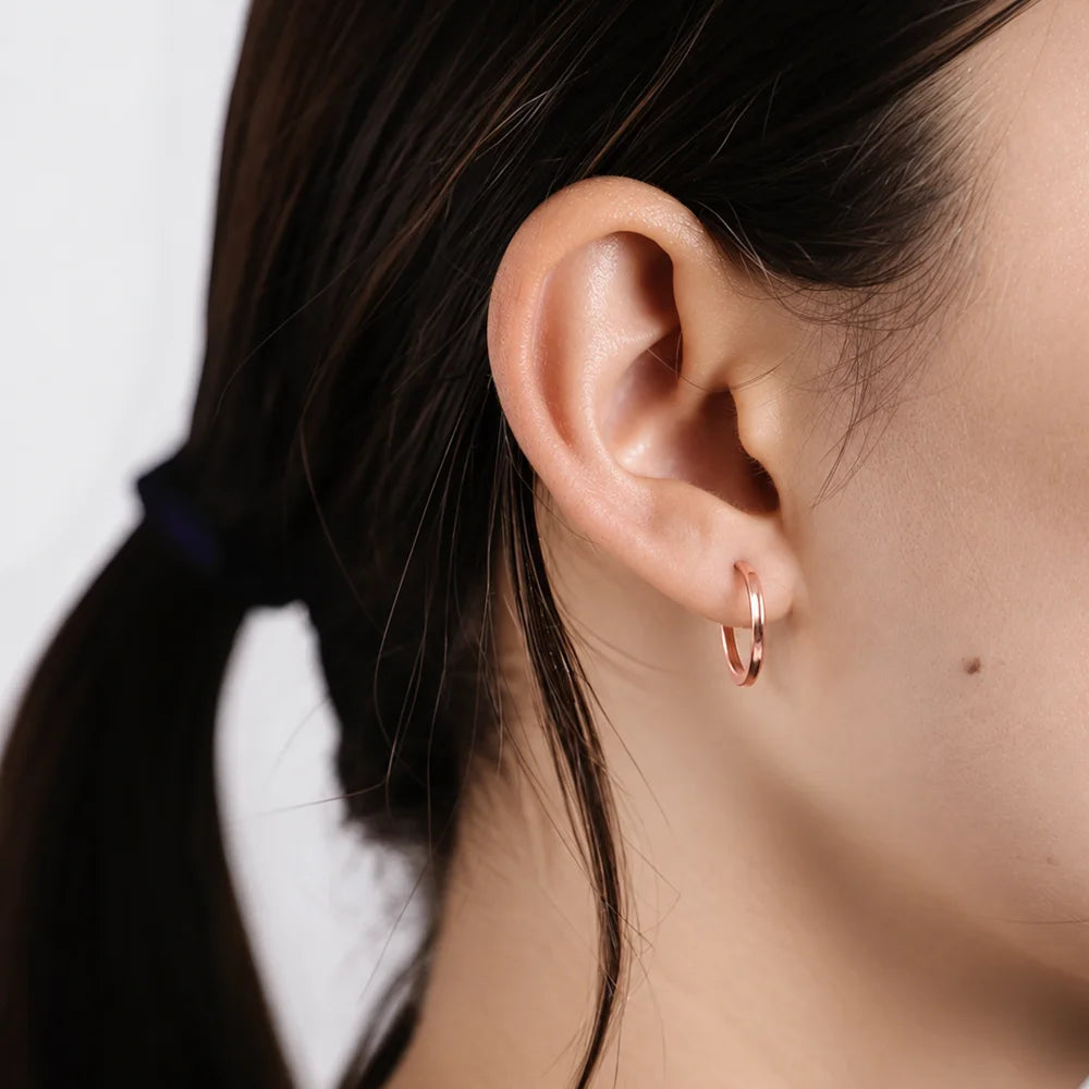 A close-up of a person's ear wearing a small rose gold hoop earring against a clean white background.