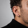 A close-up of a man's ear wearing a polished silver hoop earring, with curly dark hair, stubble, and a black turtleneck against a neutral gray background.