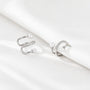 A pair of silver-toned open hoop ear cuffs with small white pearl beads and a row of clear stones, displayed on a draped white satin fabric.
