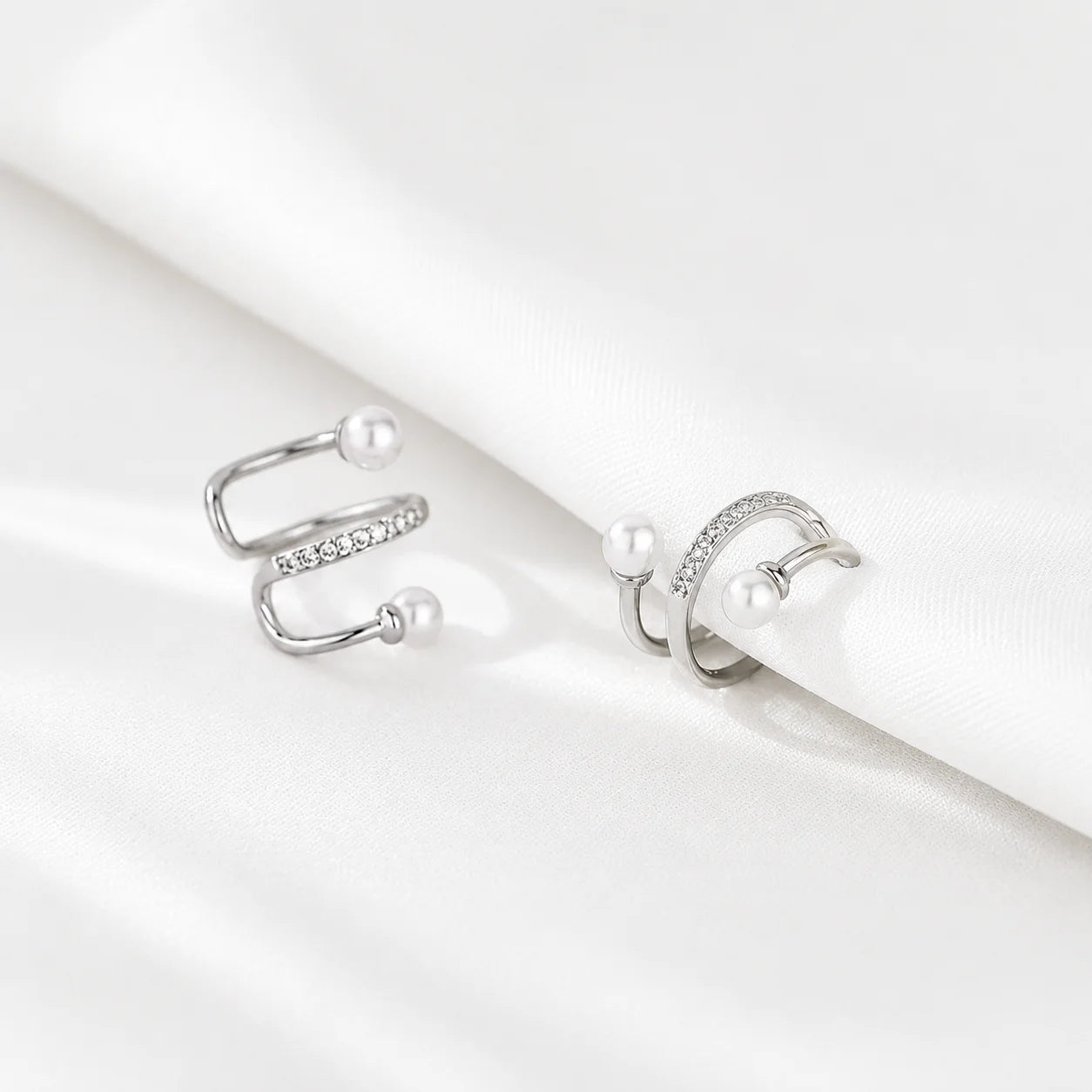 A pair of silver-toned open hoop ear cuffs with small white pearl beads and a row of clear stones, displayed on a draped white satin fabric.
