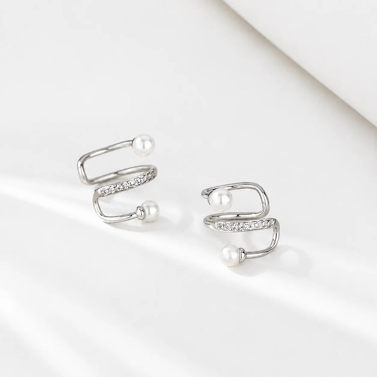A pair of silver wrap-style hoop earrings with small clear stones and white pearl beads, photographed on a clean white surface.