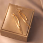 Gold ribbon dangle clip-on earrings in elegant gift box, overhead angle view on beige background
