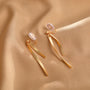Gold ribbon dangle clip-on earrings displayed on beige fabric, showing curved metal design with clear ear clips