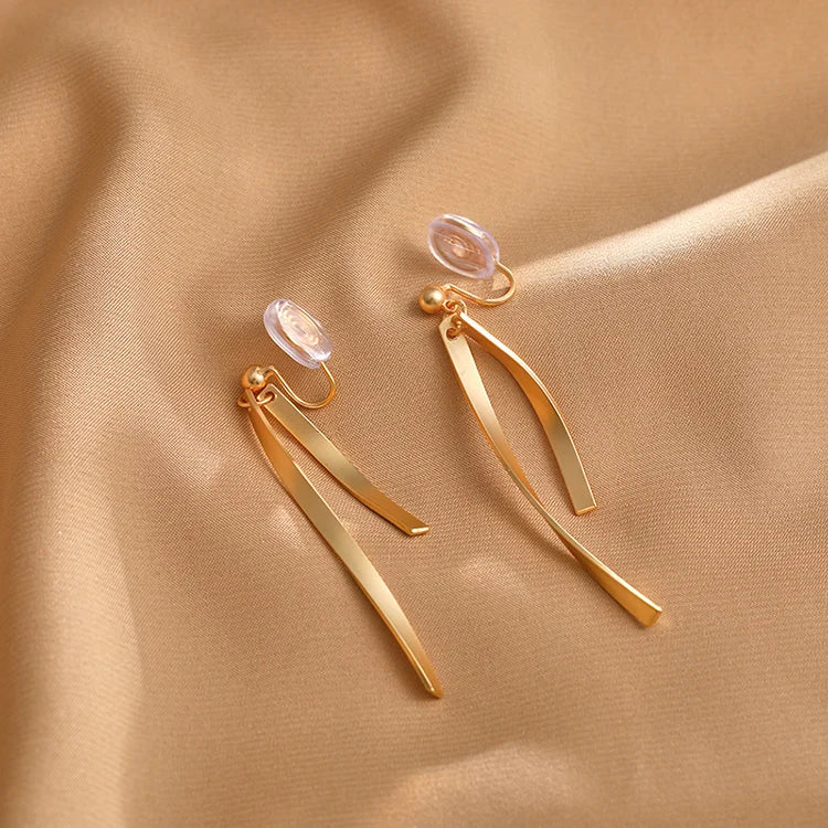 Gold ribbon dangle clip-on earrings displayed on beige fabric, showing curved metal design with clear ear clips