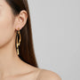 A close-up of a woman's ear wearing a pair of long, abstract gold hoop earrings against a light gray background.