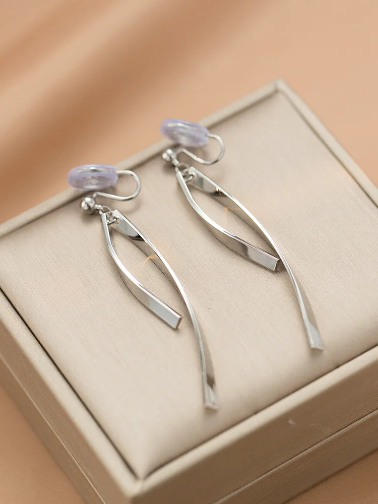 Silver ribbon dangle clip-on earrings displayed on cream jewelry box, overhead view showing sleek metallic design