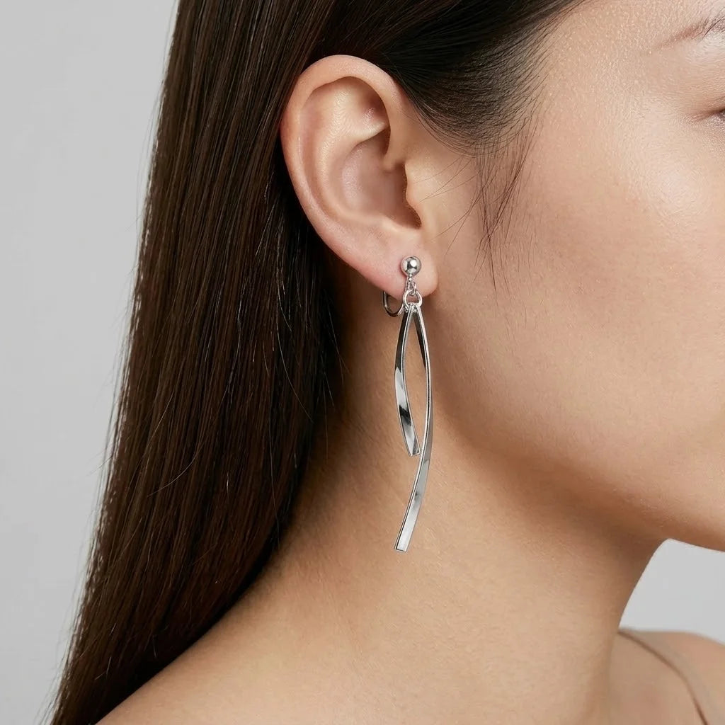 A close-up of a model's ear wearing a silver huggie hoop earring with a long, curved, polished silver dangle charm.