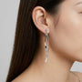 A close-up of a model's ear wearing a silver huggie hoop earring with a long, curved, polished silver dangle charm.