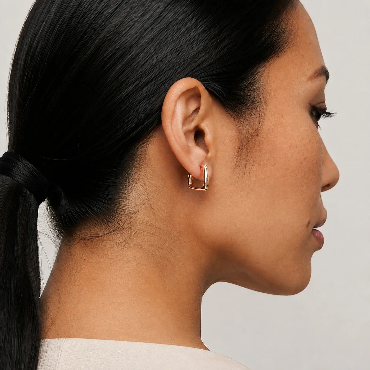 A close-up side profile of a woman with dark hair tied back, wearing a small gold square hoop earring