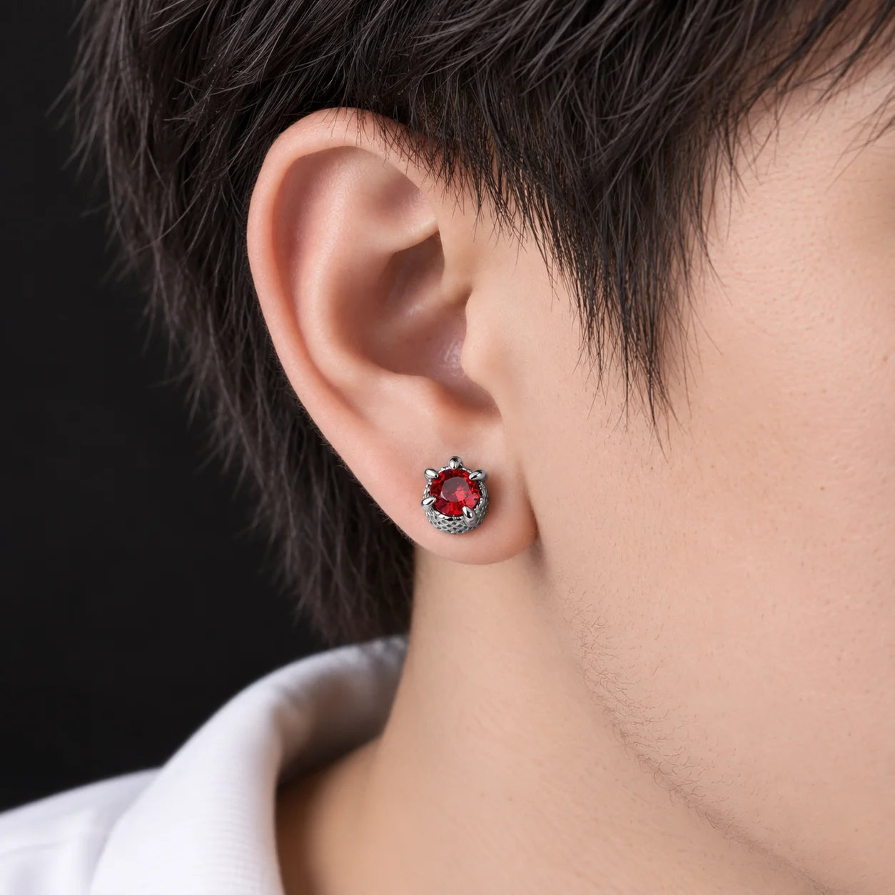 A close-up of a person's ear wearing a silver-toned stud earring with a round red gemstone surrounded by small clear stones, against a dark background.