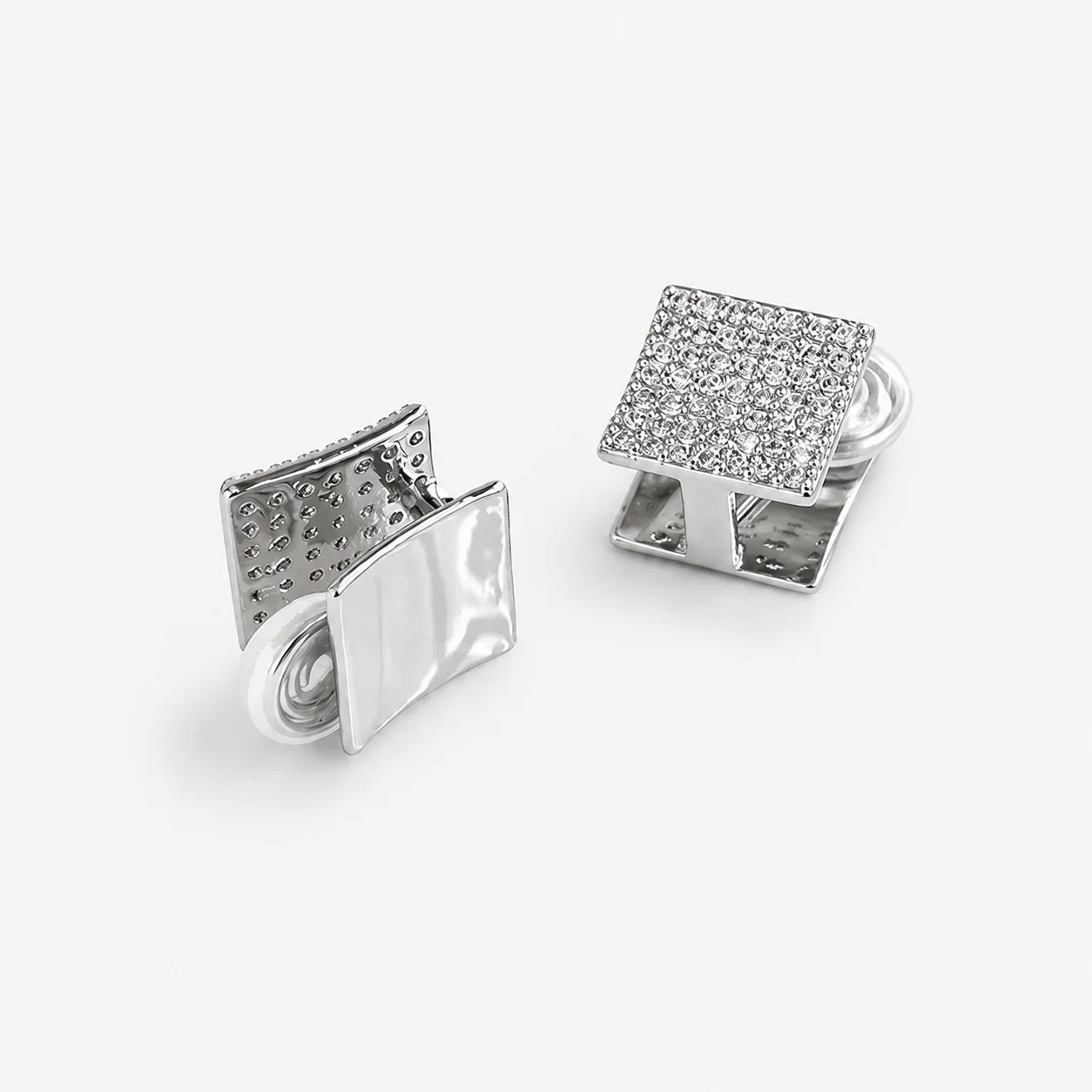 A pair of silver-toned square cufflinks on a light gray background. One cufflink is shown from the front, featuring a flat, polished metallic surface, while the other is turned to show the back, which has a textured, diamond-like pavé pattern on its top face.