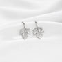 A pair of silver leaf-shaped dangle earrings displayed on a white ceramic plate against a soft white fabric background.