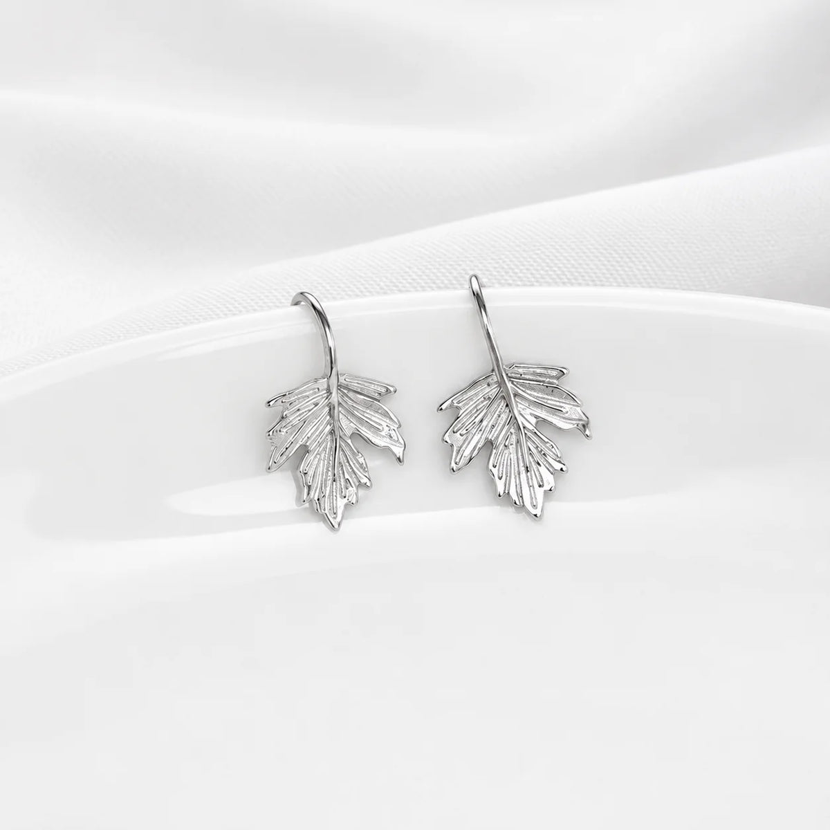 A pair of silver leaf-shaped dangle earrings displayed on a white ceramic plate against a soft white fabric background.