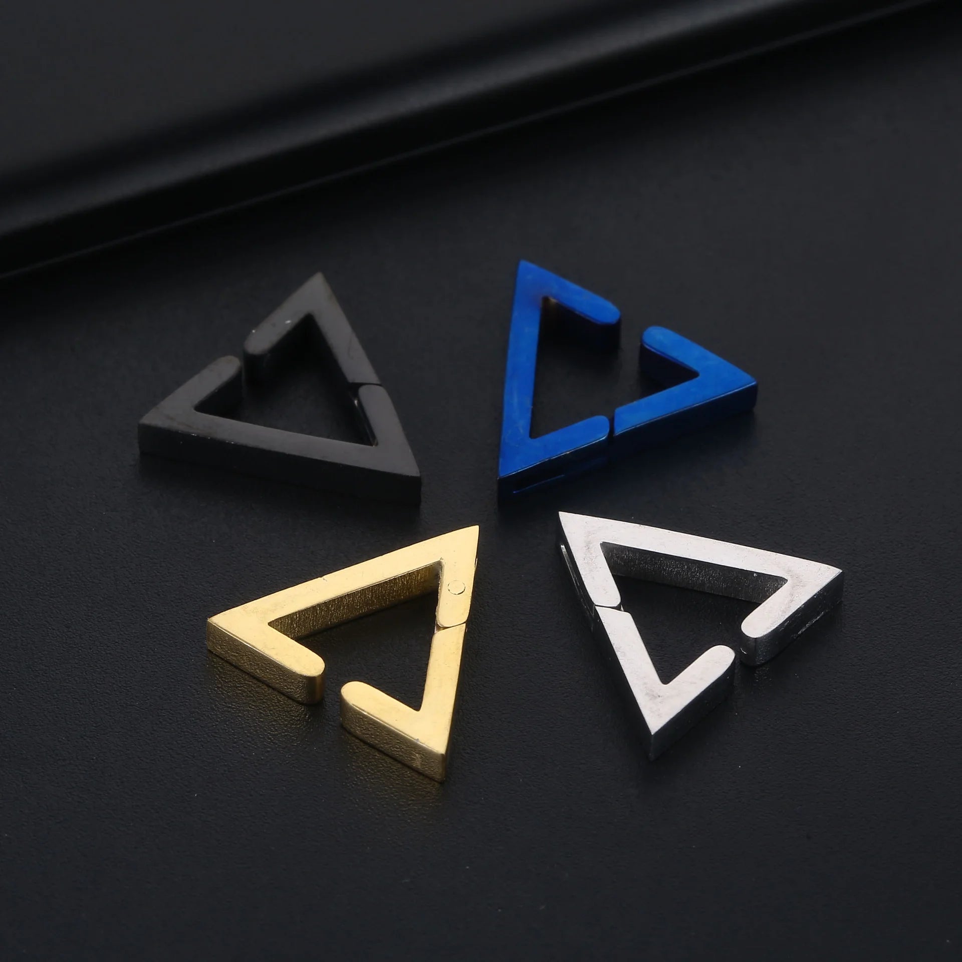 Four metallic geometric triangle clip-on earrings in gold, blue, silver and gunmetal displayed on dark surface