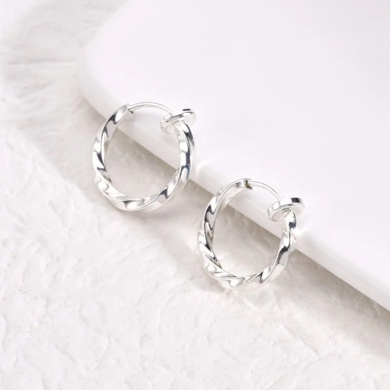 Twisted huggie clip on hoop earrings in silver displayed on white marble surface, angled view