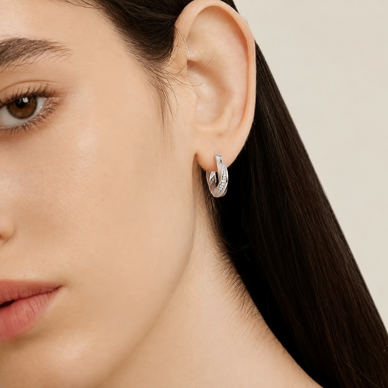 A close-up of a model's ear wearing a small silver hoop earring with a textured, diamond-like pavé surface, set against a clean, light beige background.