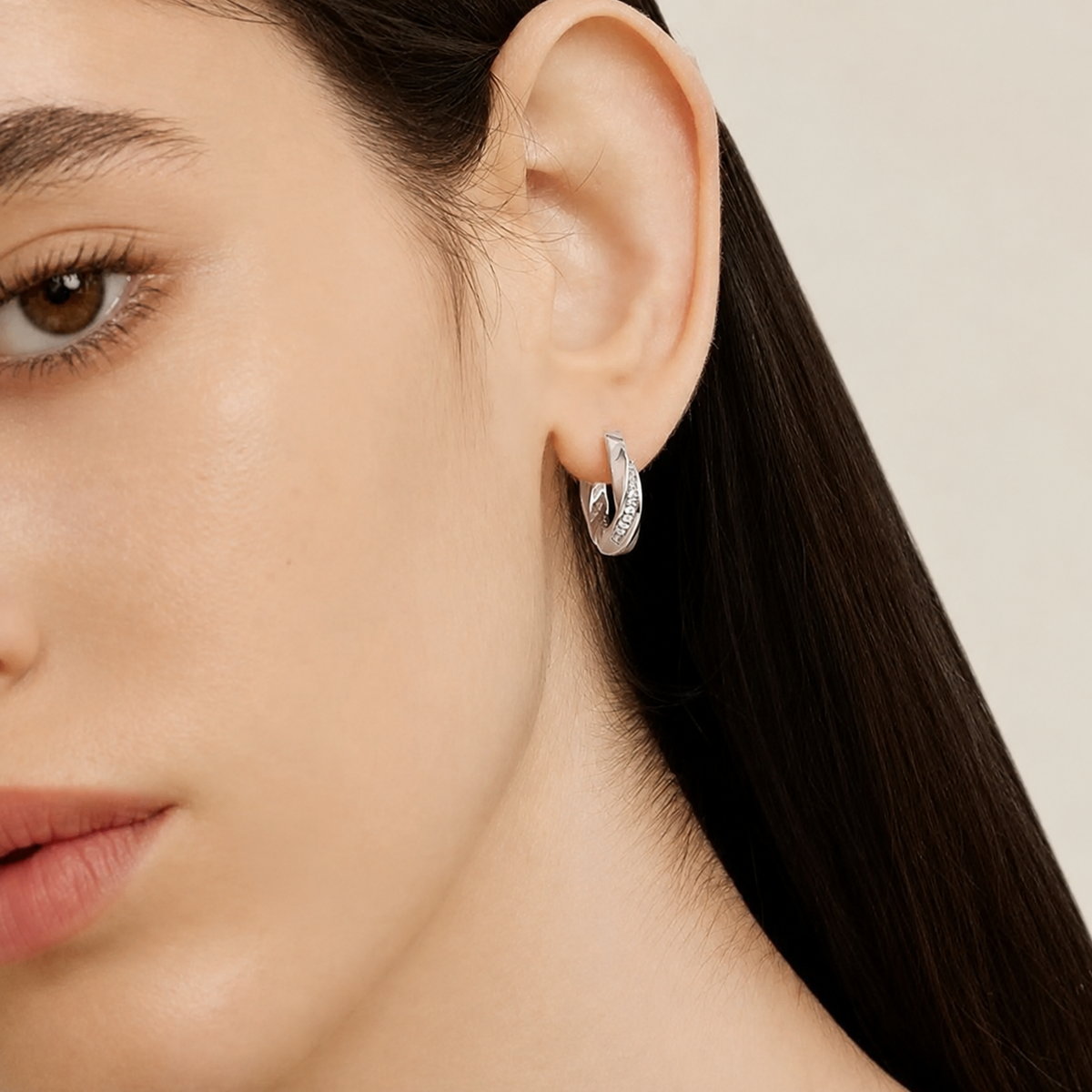 A close-up of a model's ear wearing a small silver hoop earring with a textured, diamond-like pavé surface, set against a clean, light beige background.