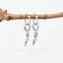 A pair of silver huggie hoop earrings with dangling twisted branch-like charms, hanging from a natural brown twig against a clean white background.