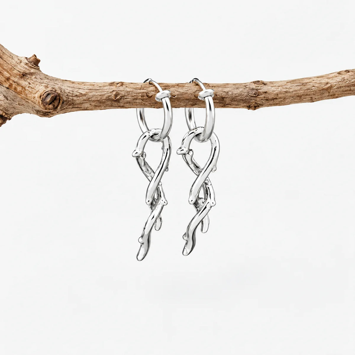 A pair of silver huggie hoop earrings with dangling twisted branch-like charms, hanging from a natural brown twig against a clean white background.