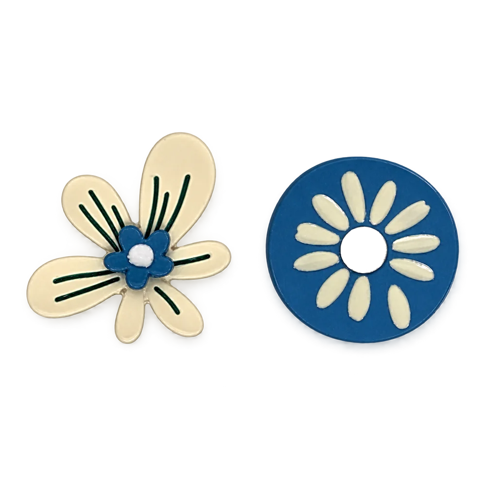 A pair of floral clip on earrings a white background. One is featuring a cream-colored five-petal flower with a blue center, white dot, and green leaf-like lines.