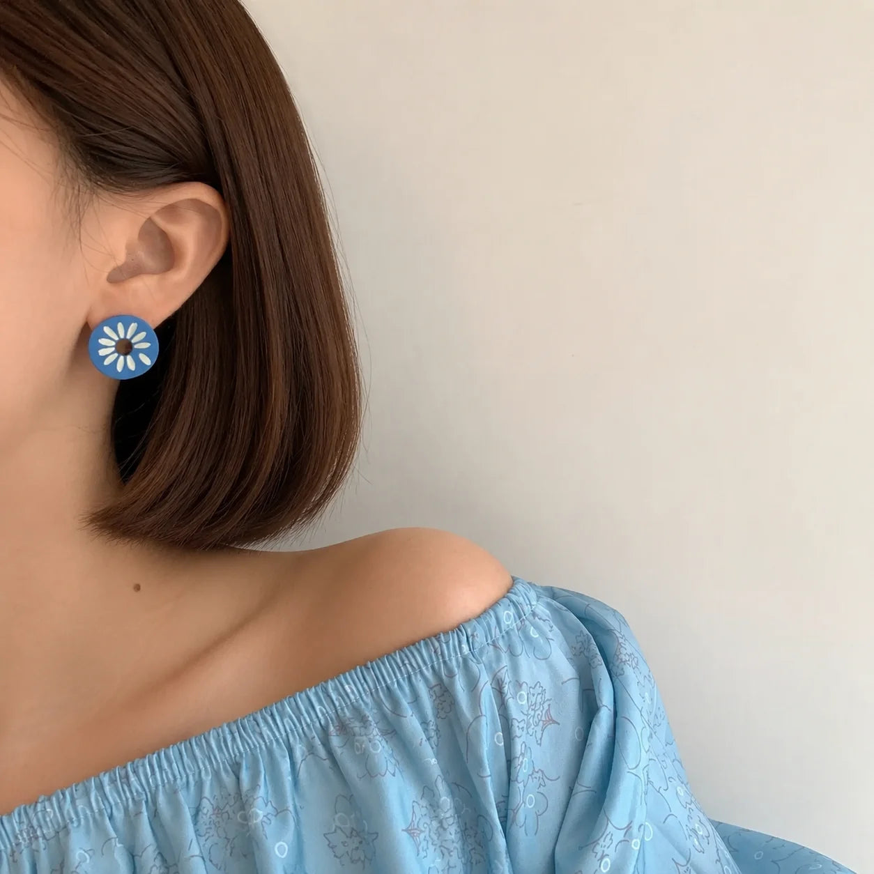 A close-up of a woman with short brown hair wearing a blue and white daisy-shaped stud earring, paired with a light blue off-the-shoulder top against a plain off-white wall.