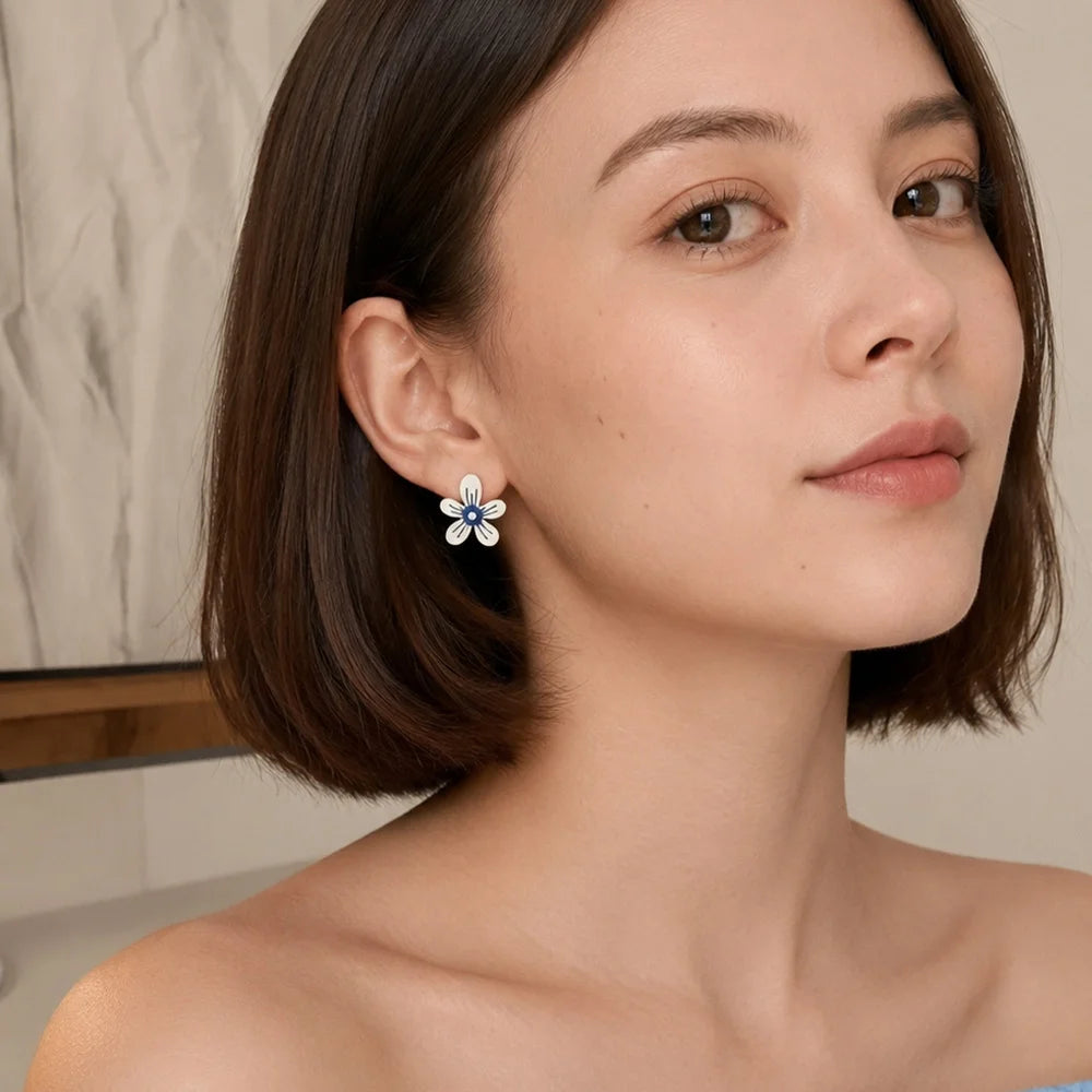 A close-up of a woman with short brown hair wearing a small flower-shaped stud earring with white petals and a blue center, set against a softly blurred indoor background.
