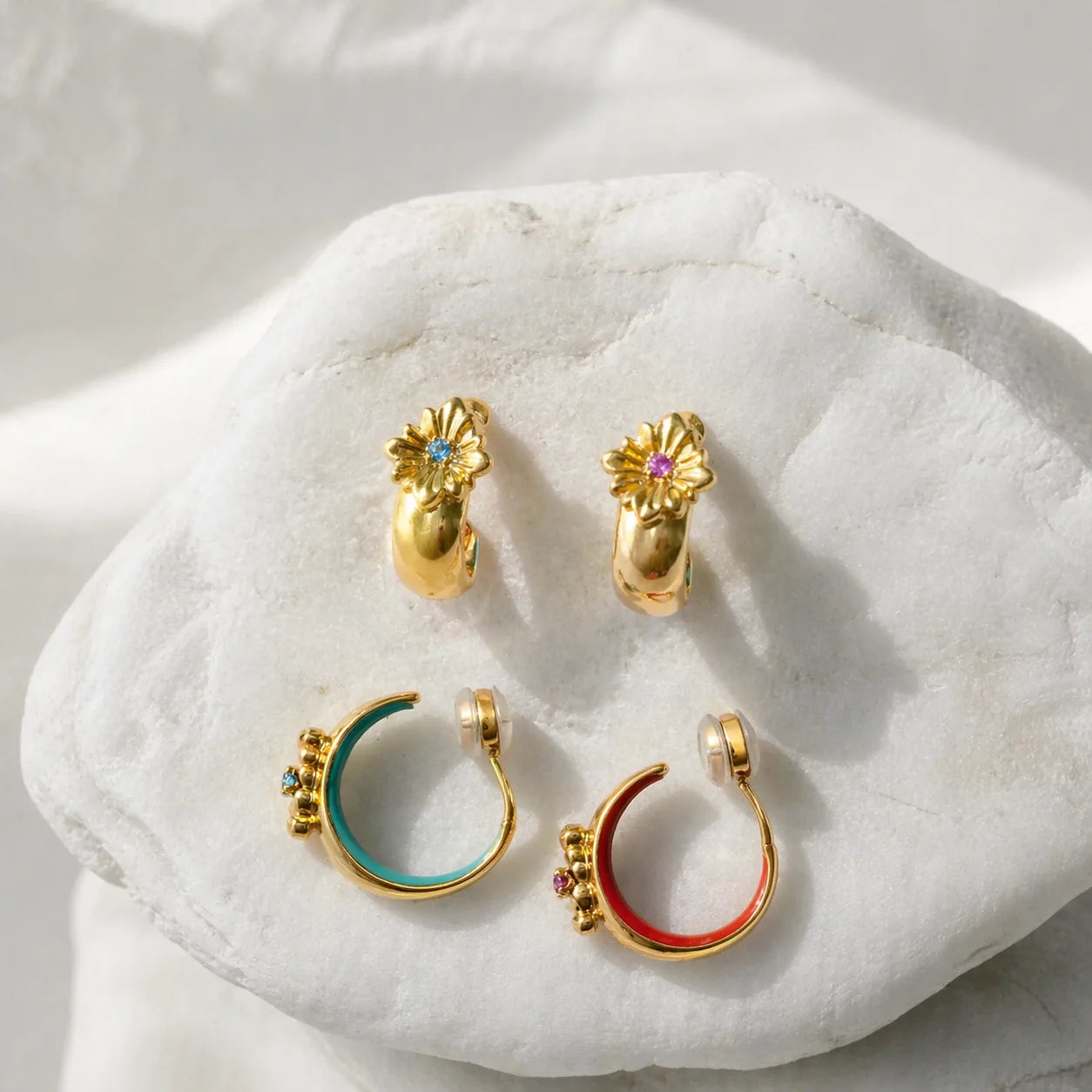A collection of gold bohemian cross motif clip on hoops with turquoise and crimson enamel linings displayed in a lifestyle setting.