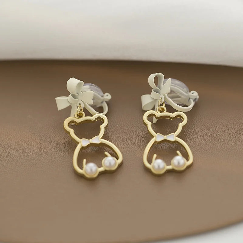 Gold bow-tied bear pearl clip-on earrings displayed on beige surface, front view showing white bow tops and pearl accents