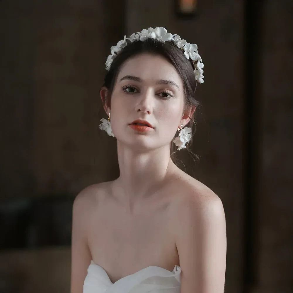 Close-up portrait of a bride in a strapless dress wearing a white ceramic flower headband and the matching cascading blossom clip-on earrings, facing forward.