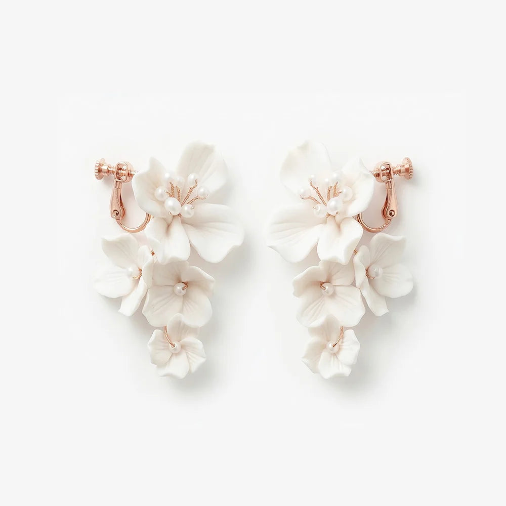Flat lay front view of a matching pair of handmade white ceramic flower cascading clip-on bridal earrings with pearl centers on rose gold hardware, isolated on a white background.