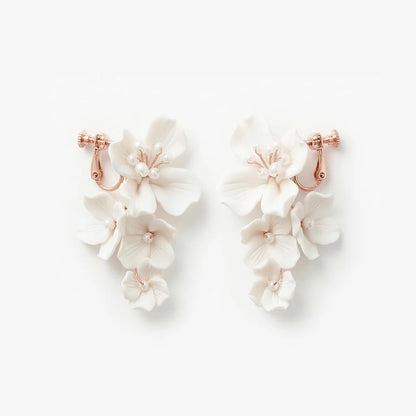 Flat lay front view of a matching pair of handmade white ceramic flower cascading clip-on bridal earrings with pearl centers on rose gold hardware, isolated on a white background.