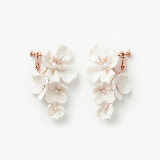 Flat lay front view of a matching pair of handmade white ceramic flower cascading clip-on bridal earrings with pearl centers on rose gold hardware, isolated on a white background.
