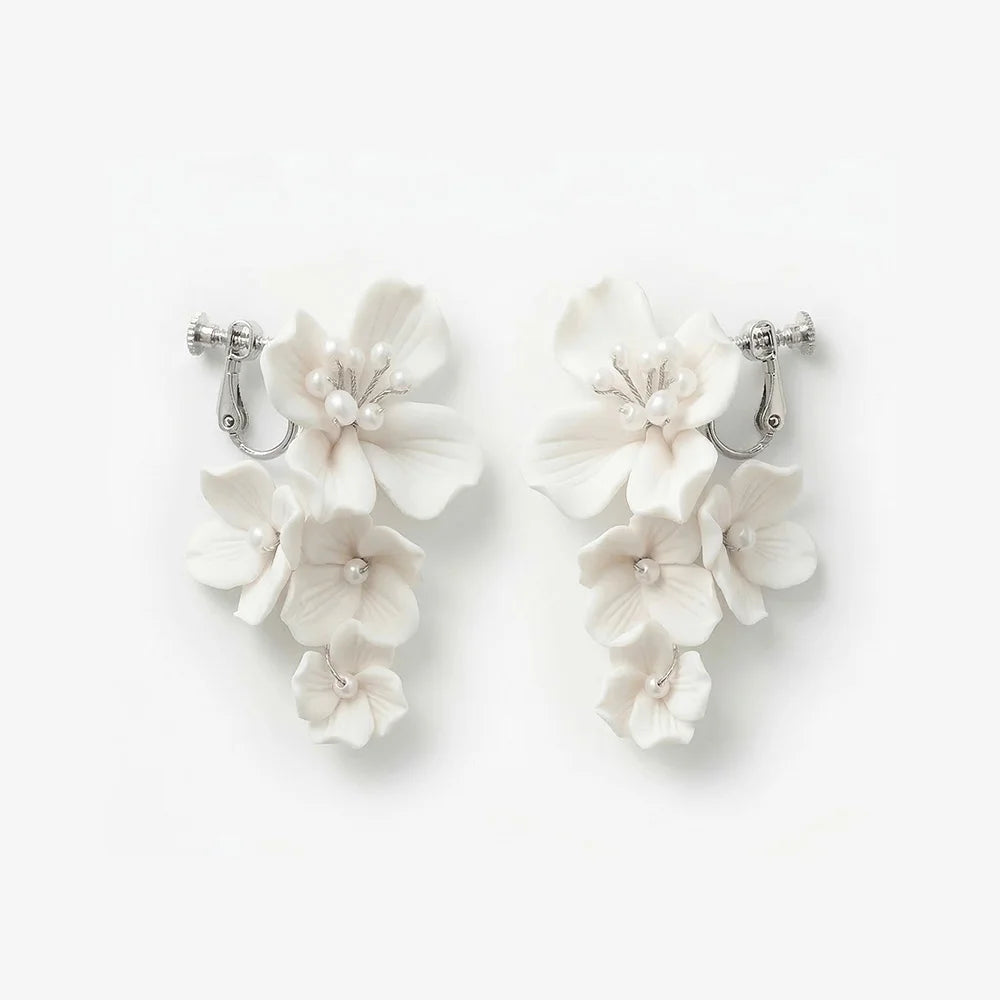 Flat lay front view of a matching pair of cascading handmade white ceramic flower clip-on bridal earrings with pearl clusters on polished silver-plated hardware, isolated on a clean white background.