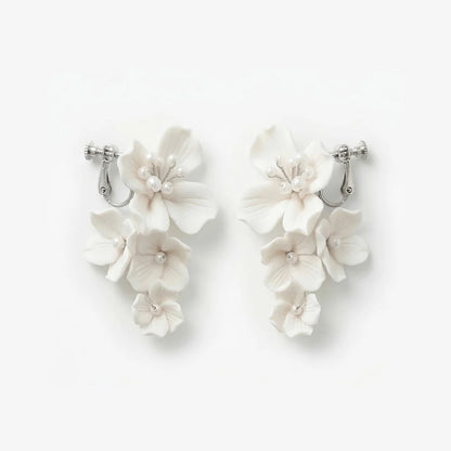 Flat lay front view of a matching pair of cascading handmade white ceramic flower clip-on bridal earrings with pearl clusters on polished silver-plated hardware, isolated on a clean white background.