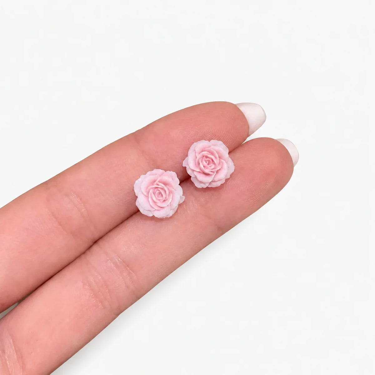 A pair of small light pink rose stud earrings resting on a person's fingers against a plain white background.