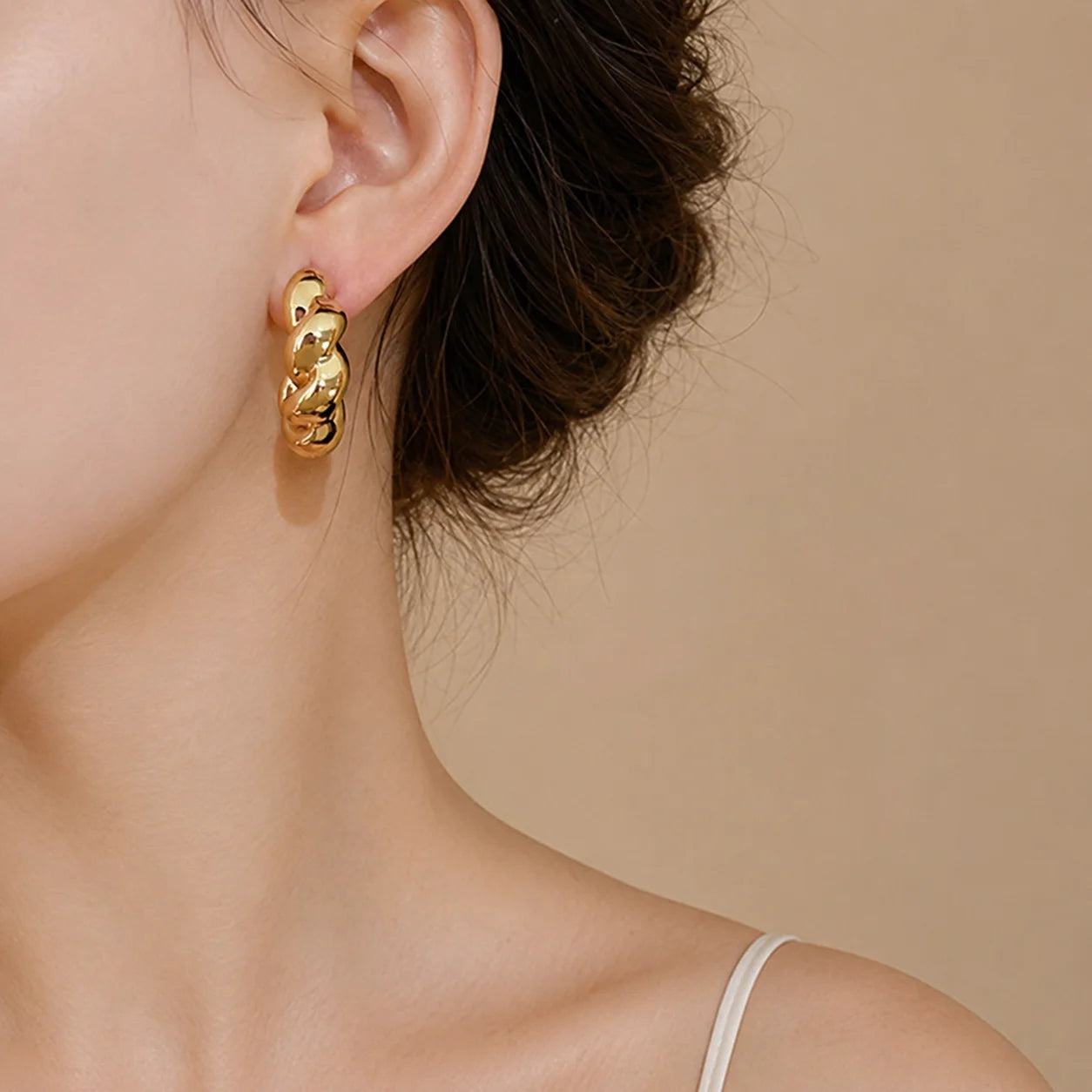 A close-up of a model's ear wearing a chunky, textured gold hoop earring against a warm beige background.
