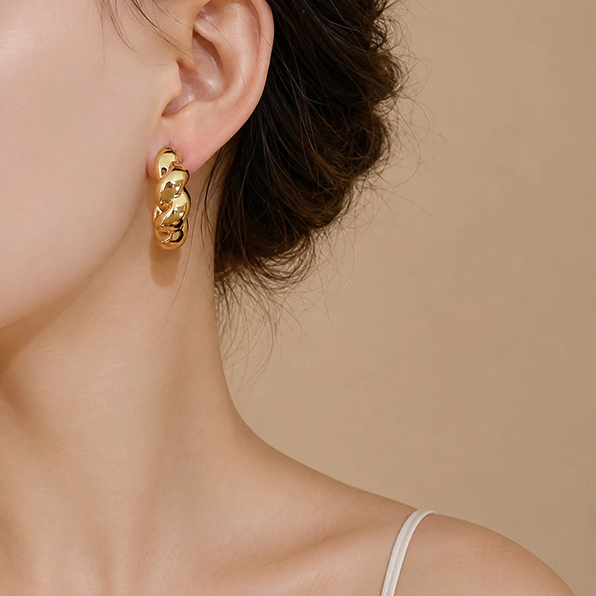 A close-up of a model's ear wearing a chunky, textured gold hoop earring against a warm beige background.
