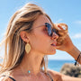 A woman outdoors by the ocean wearing gold leaf-shaped drop earrings, black cat-eye sunglasses, and layered gold necklaces.
