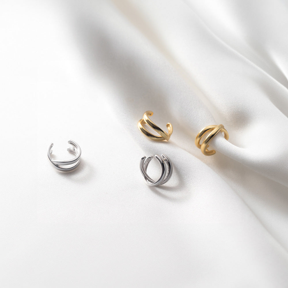Une collection de manchettes d'oreilles Helix Cross, disposées sur un tissu blanc doux, met en valeur les finitions or et argent. Les courbes élégantes de ces manchettes épousent les lignes naturelles de l'oreille, ce qui en fait un choix polyvalent pour le bureau ou pour apporter une touche raffinée à une tenue décontractée chic.