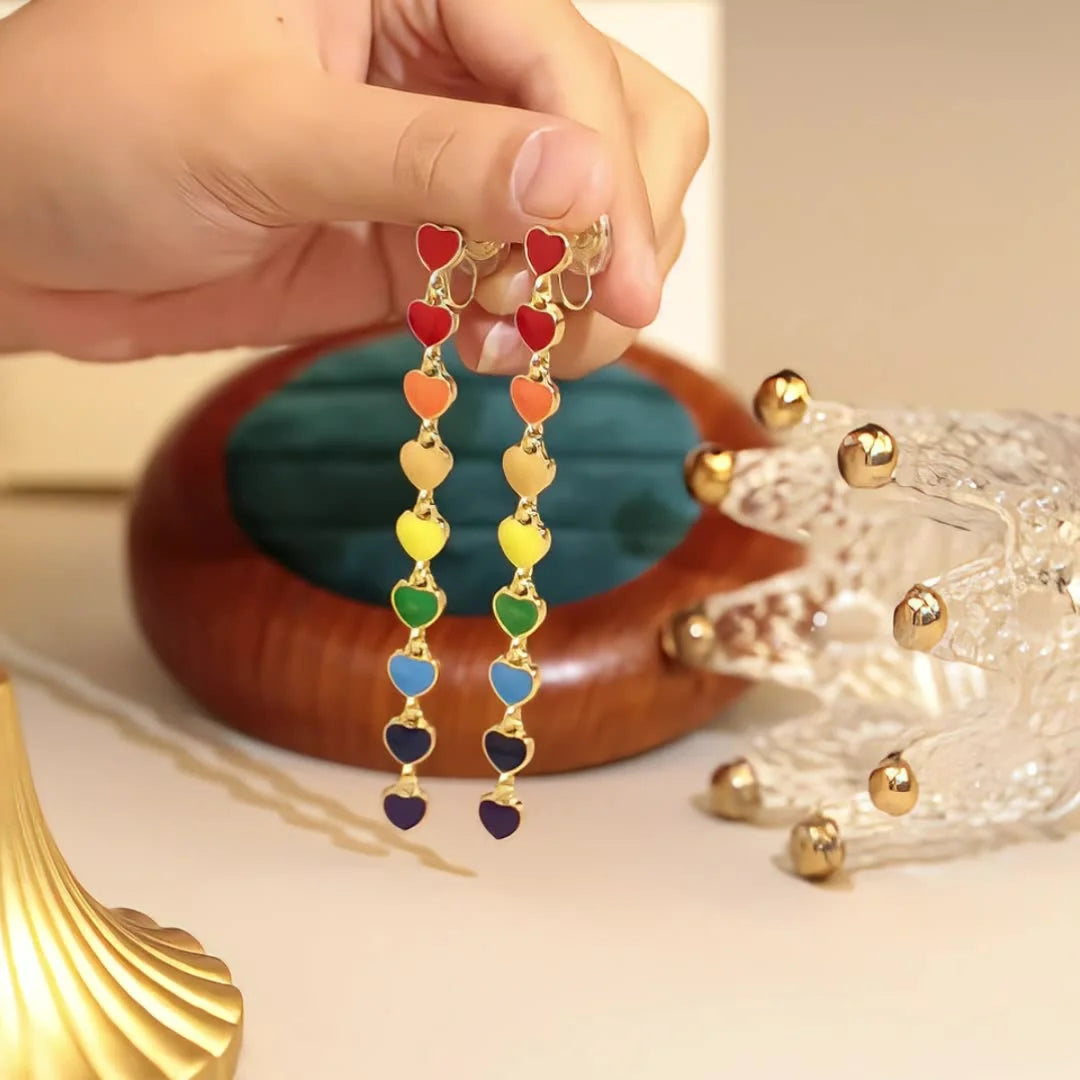 Rainbow Heart Cascade clip-on earrings held in hand, featuring colorful enamel hearts in red, orange, yellow, green, blue and purple on gold-tone chain links
