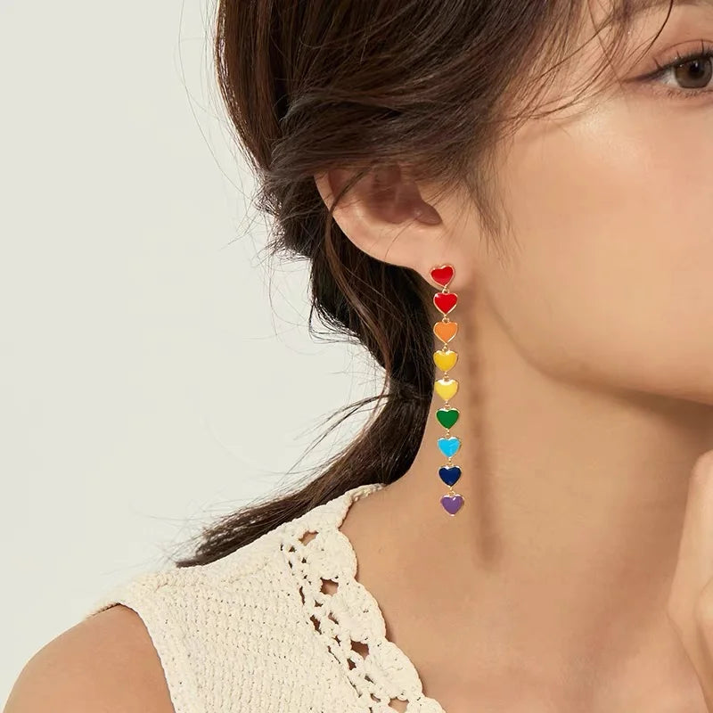 A close-up of a woman wearing a long dangling earring made of stacked rainbow-colored hearts, with her ear, hair, and shoulder visible against a light neutral background.
