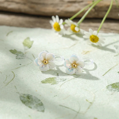 A pair of white iridescent flower clip-on earrings with a sculptural 3D petal design displayed on a white textured surface.