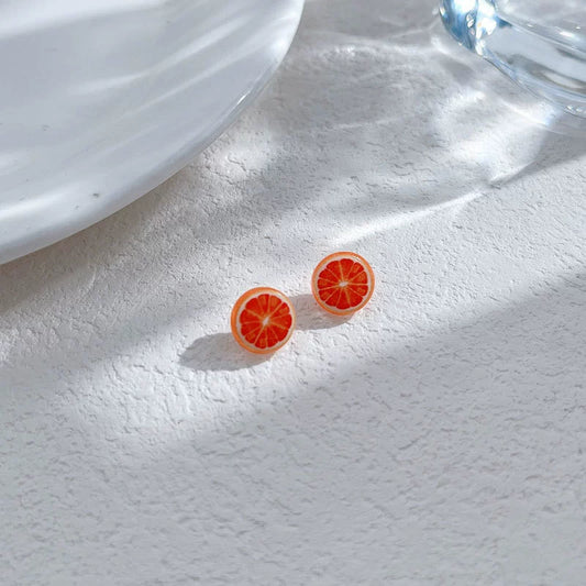 Two Zesty Grapefruit Magnetic Stud Earrings displayed on a textured white surface, with dappled sunlight, highlighting their vibrant color.