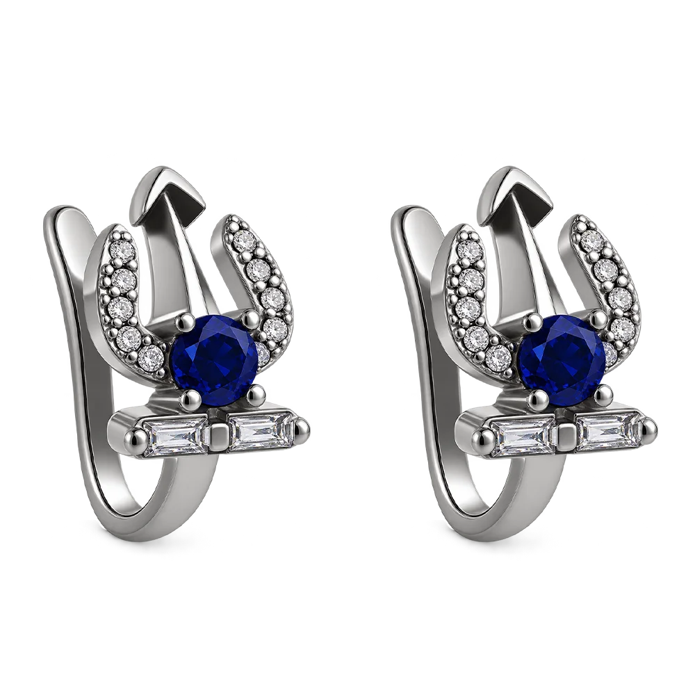 A pair of silver-toned huggie hoop earrings featuring a central round-cut blue sapphire, surrounded by small clear cubic zirconia stones in a crescent shape, with a small rectangular bar of baguette-cut clear stones at the bottom, shown on a plain white background.
