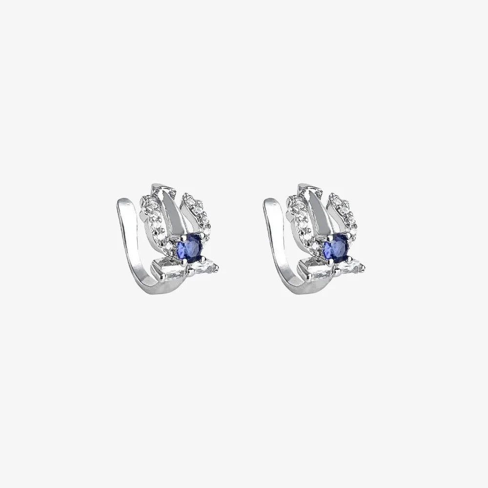 A pair of silver trident clip-on ear cuffs, each adorned with a sapphire blue cubic zirconia and white accent stones, on a white background.