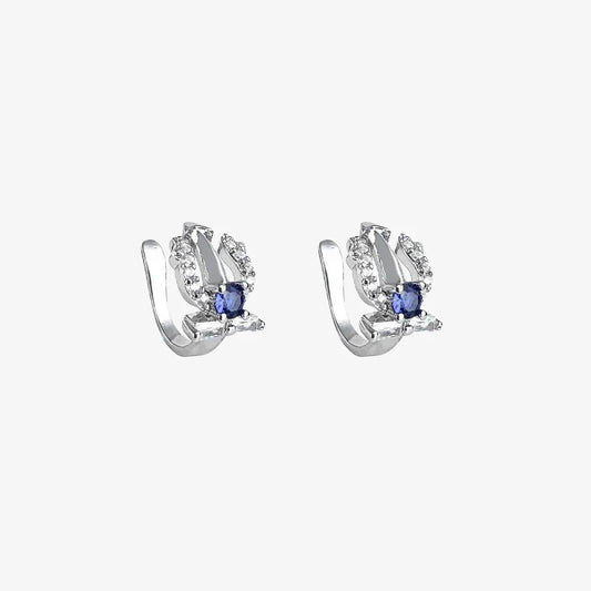 A pair of silver trident clip-on ear cuffs, each adorned with a sapphire blue cubic zirconia and white accent stones, on a white background.