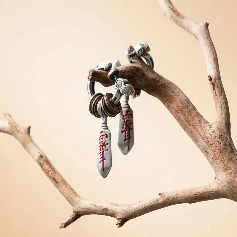 Two matching Katana dagger single clip-on earrings are artfully displayed, hanging from a dry, natural wooden branch against a neutral cream background, suggesting a natural and artisanal feel.