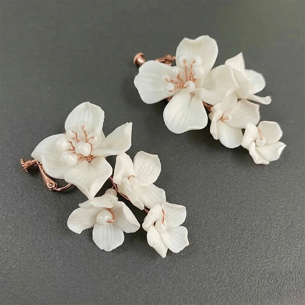 Diagonal top-down view of the pair of handmade porcelain-style white flower cascading clip-on earrings resting on a dark, textured charcoal surface, showing detail of the multi-tiered blooms and screw-back closures.