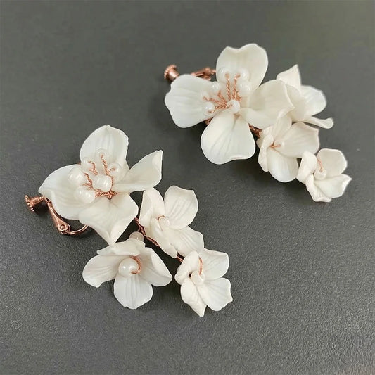 Diagonal top-down view of the pair of handmade porcelain-style white flower cascading clip-on earrings resting on a dark, textured charcoal surface, showing detail of the multi-tiered blooms and screw-back closures.