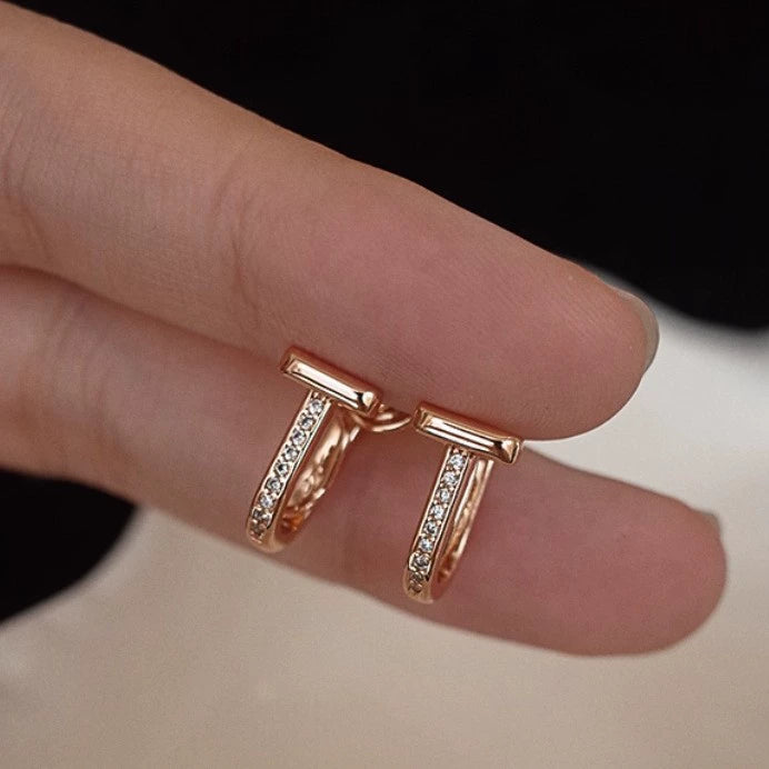Rose gold earrings with small diamonds T-motif design on a finger against a neutral background